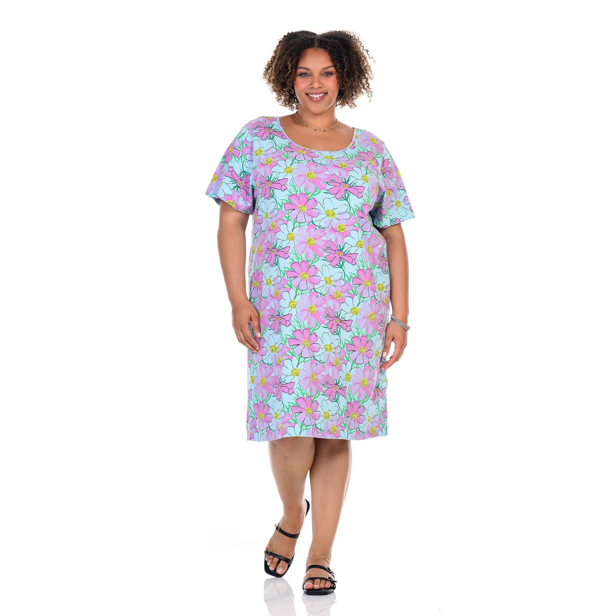A woman stands smiling against a white background, wearing the Fresh Produce Clothes Spring Day Sanibel Dress | Plus Size—a short-sleeve, knee-length cotton dress with a vibrant floral print featuring pink, green, and blue accents. She pairs the look with black sandals and minimal jewelry. This plus size dress offers both comfort and style for any occasion.