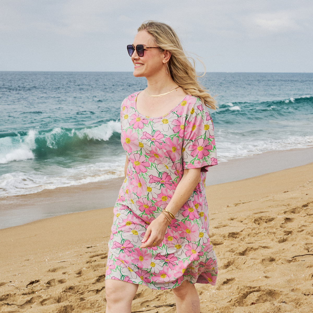 A woman, smiling as she walks along a sandy beach near the ocean, wears the Spring Day Sanibel Dress | Plus Size by Fresh Produce Clothes and sunglasses. Waves crash in the background under a partly cloudy sky.