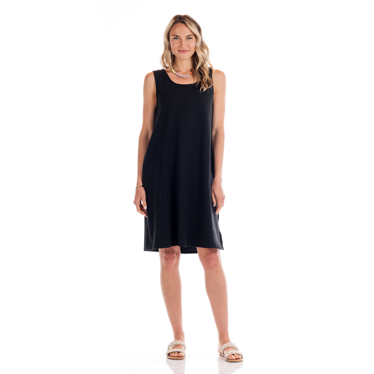 A woman stands facing forward, wearing the Fresh Produce Clothes Sun Dress—a sleeveless, black cotton jersey knee-length dress—paired with sandals. With wavy blonde hair and a smile, she poses against a plain white background. This versatile sun dress is proudly made in the USA.