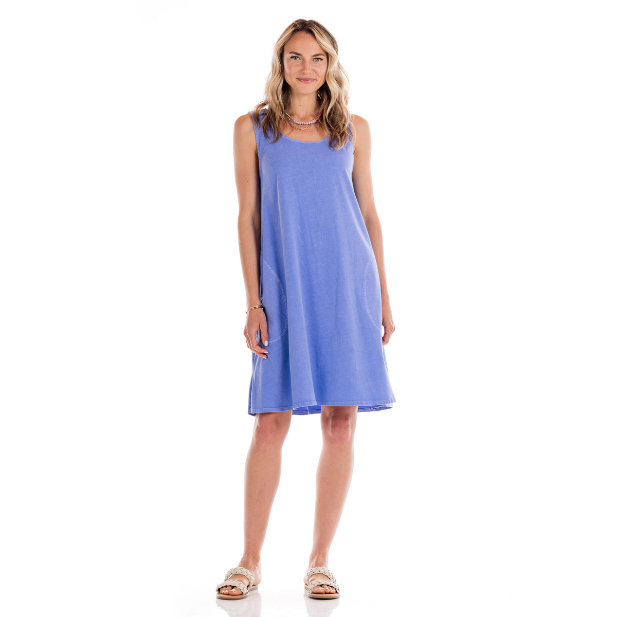 A woman stands smiling against a white background, wearing the Fresh Produce Clothes Sun Dress—a sleeveless, knee-length, light blue cotton jersey sundress—paired with white sandals. Made in USA.