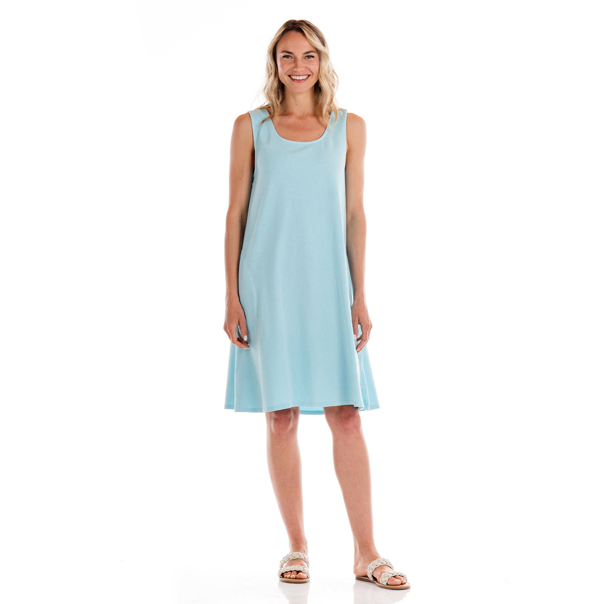 A woman stands smiling in a sleeveless, light blue cotton jersey knee-length Sun Dress by Fresh Produce Clothes with sandals, posed against a white background.