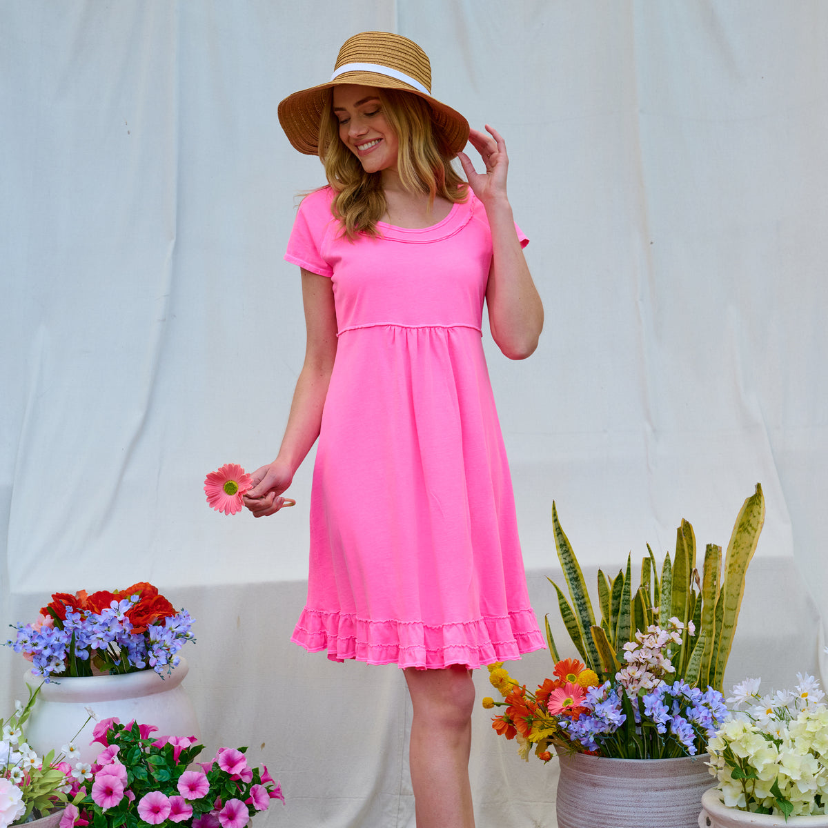 A woman wearing the Sunshine Dress by Fresh Produce Clothes and a straw hat stands among colorful potted flowers, holding a pink daisy and smiling in front of a white backdrop.