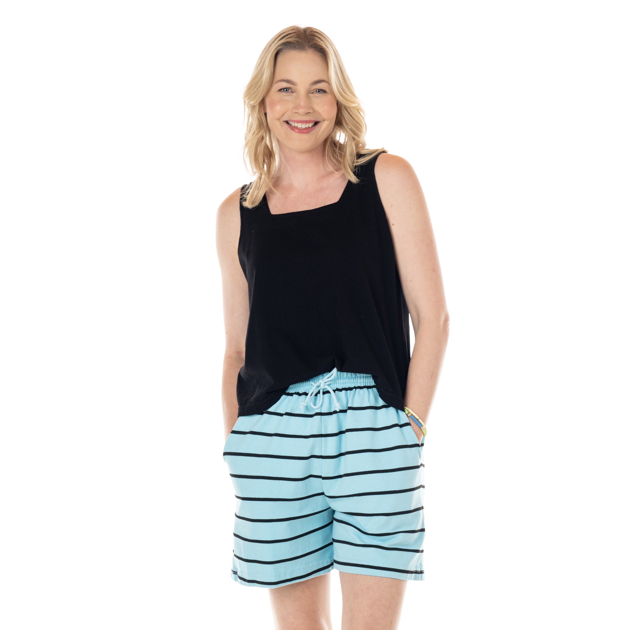 A smiling woman with blonde hair wears a black sleeveless top and Surfline Jersey Shorts by Fresh Produce Clothes—light blue women’s cotton shorts with black horizontal stripes. She stands with her hands in her pockets against a plain white background.