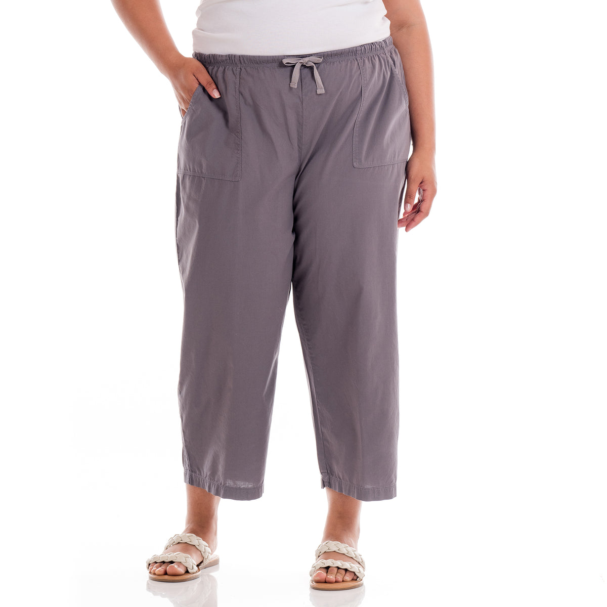 A person stands against a white background with one hand in their pocket, wearing the Fresh Produce Clothes Topstitch Sheeting Capris in gray, featuring an elastic waistband and drawstring, paired with a white top and white woven sandals.