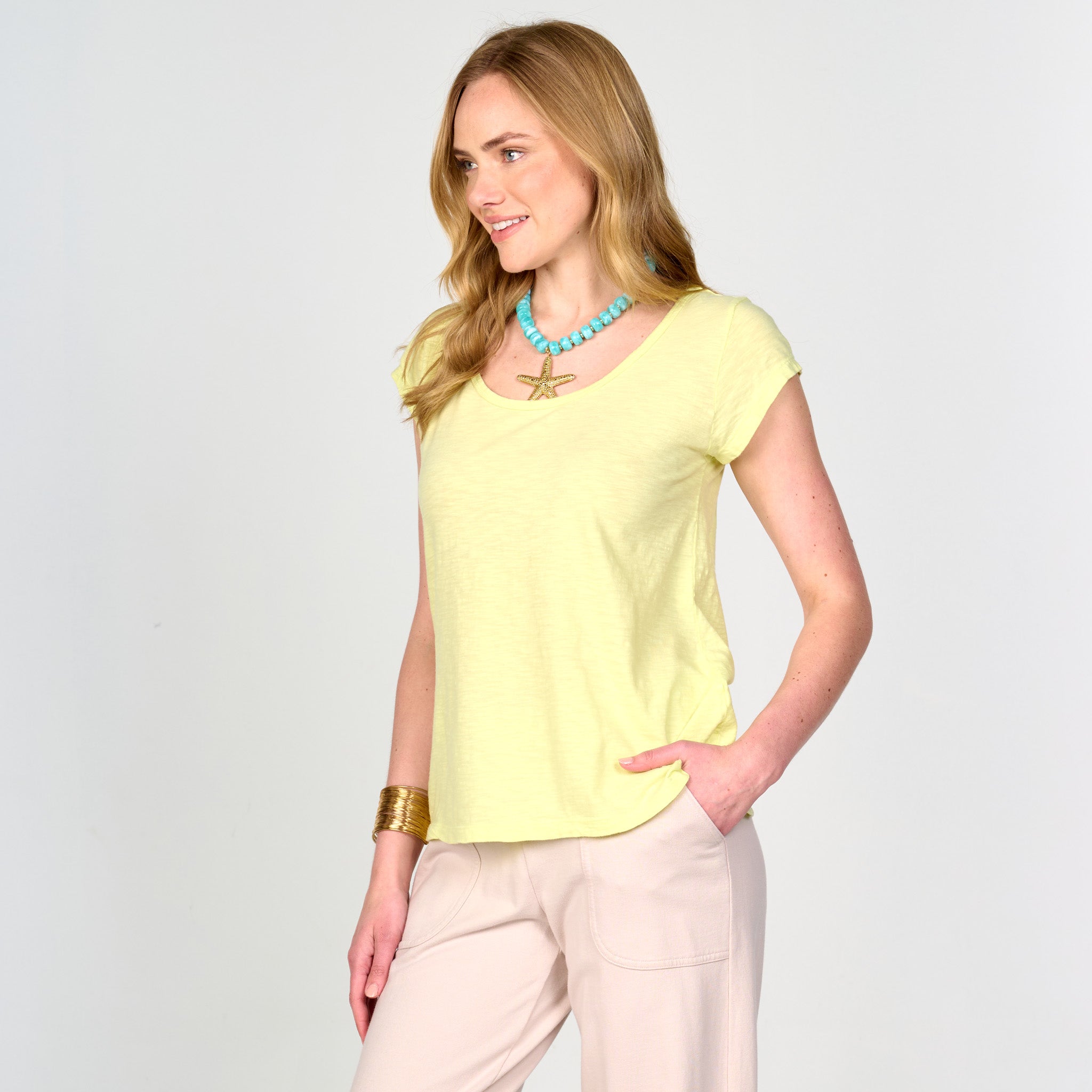 A woman with long blonde hair is wearing the Fresh Produce Clothes Vintage Scoop Neck T in yellow (Clearance), featuring cap sleeves and a slimming silhouette that flatters her figure. She pairs it with light pink pants, a turquoise star pendant necklace, and a gold bracelet, standing with one hand in her pocket and smiling, radiating cheerful style.