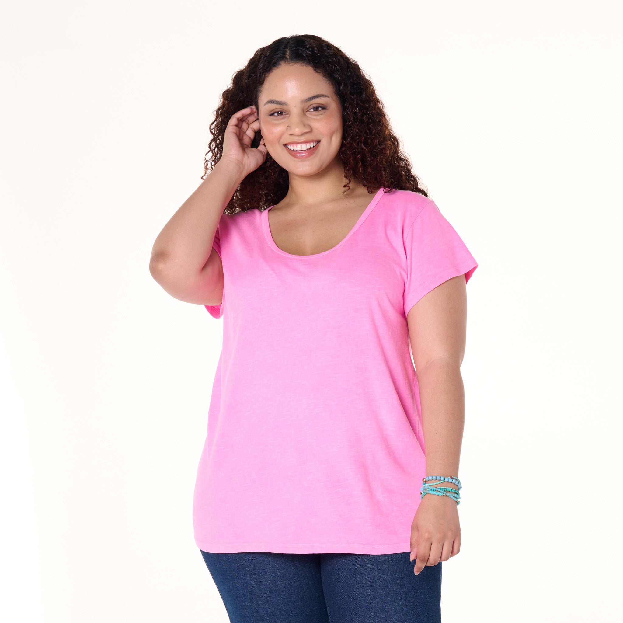 A woman with curly hair smiles, wearing the Fresh Produce Clothes Vintage Scoop Neck T in bright pink and dark blue jeans. She stands against a plain white background, one hand touching her hair and the other by her side, highlighting the slimming shape of this plus size clearance top.