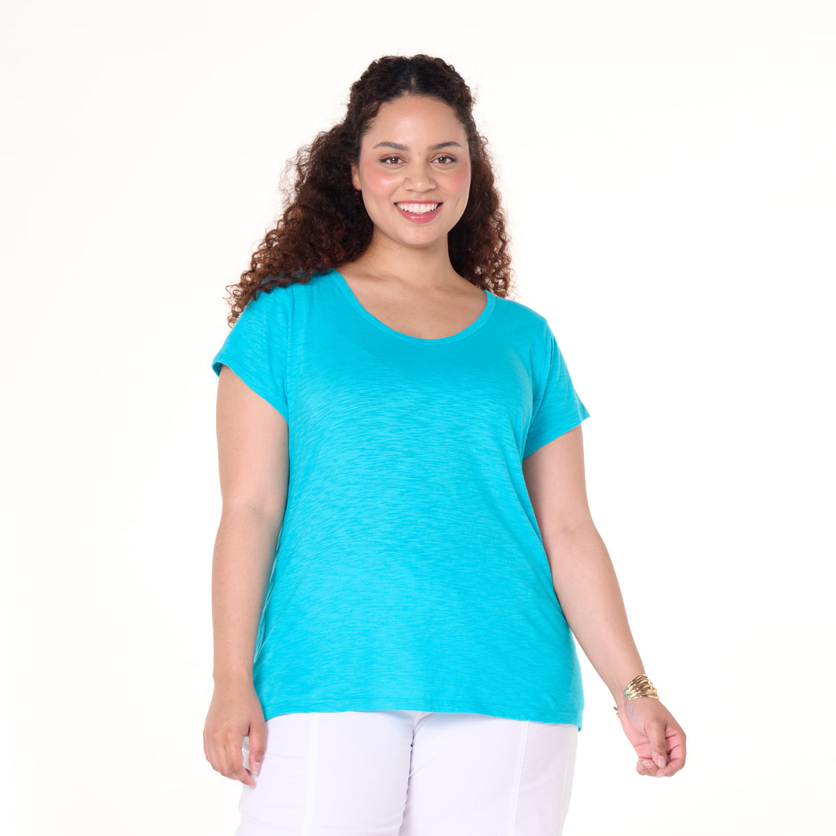 A woman with curly hair wears a bright blue Fresh Produce Clothes Vintage Scoop Neck T (Plus Size, Clearance), featuring a lightweight, slimming shape, paired with white pants. She smiles while standing against a plain white background.
