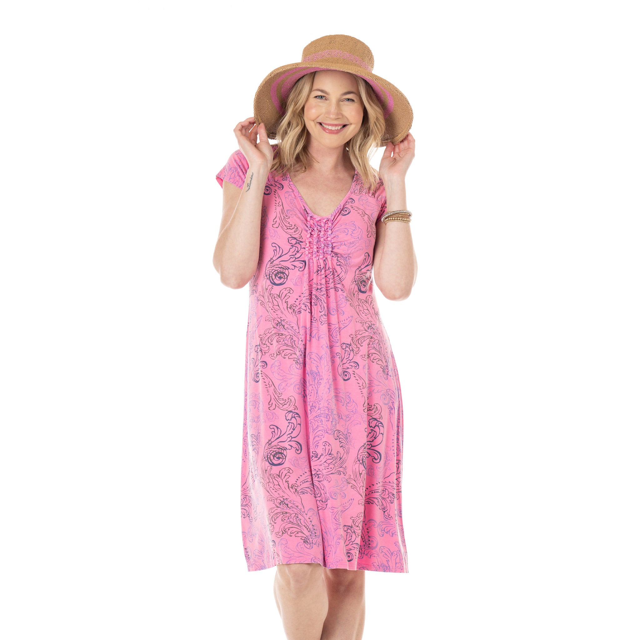 A woman wearing the Waverley Swirl Emma Dress by Fresh Produce Clothes—a pink patterned A-line dress—smiles and holds the brim of her wide-brimmed straw hat with both hands while standing against a white background.
