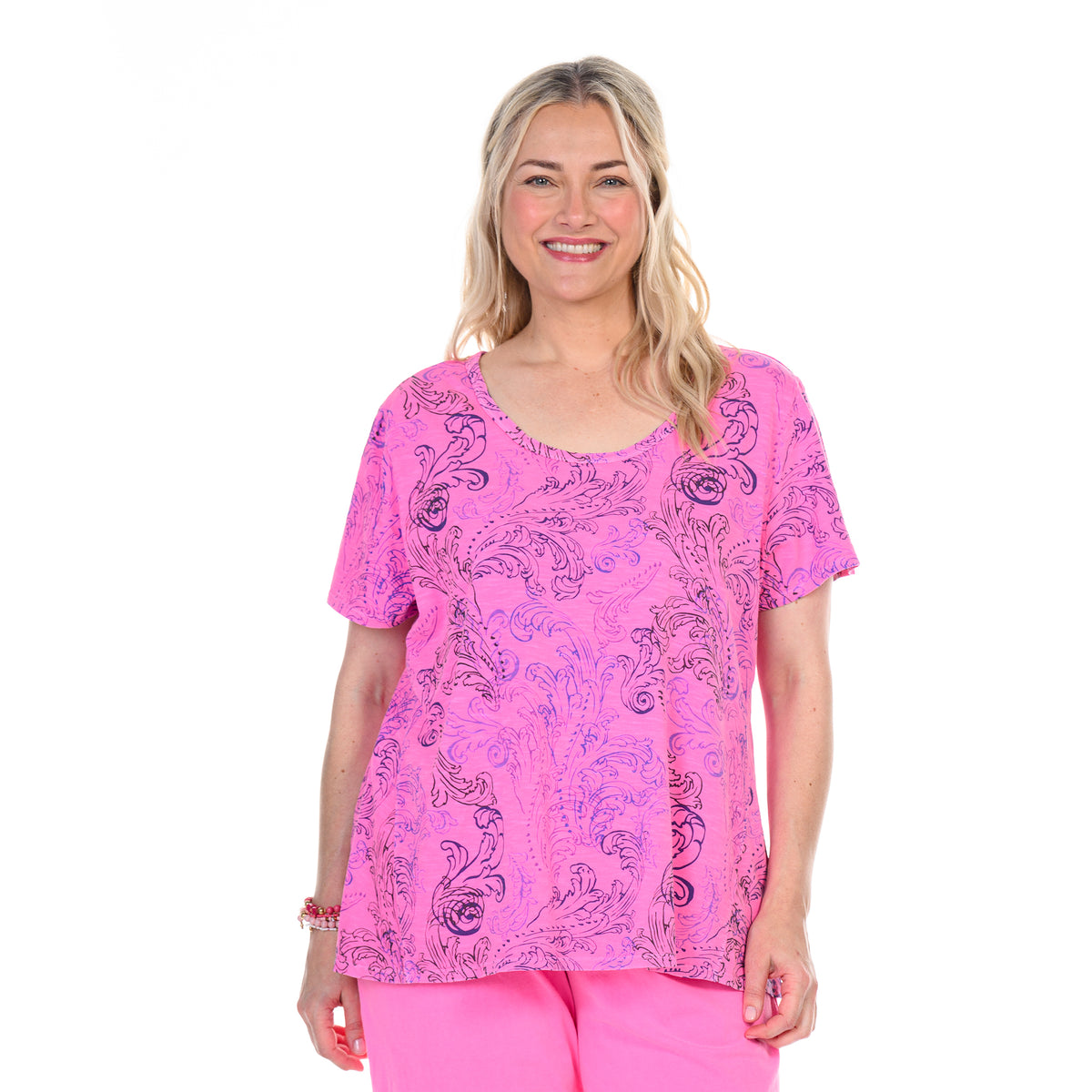 Smiling woman with long blonde hair wears the Fresh Produce Clothes Waverley Swirl Luna Top in plus size—a bright pink, flowy short-sleeve tee crafted from soft cotton slub with a black floral pattern—paired with matching pink pants, standing against a white background.