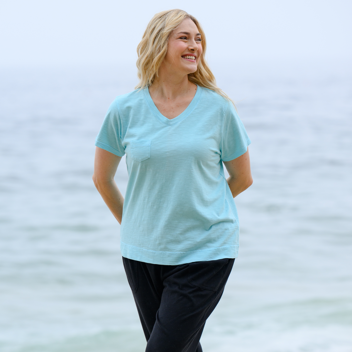 A plus size person with long blonde hair, wearing the Fresh Produce Clothes Heritage Logo Fem T | Plus Size in light blue and black pants, stands on a beach and smiles, with the ocean blurred in the background.