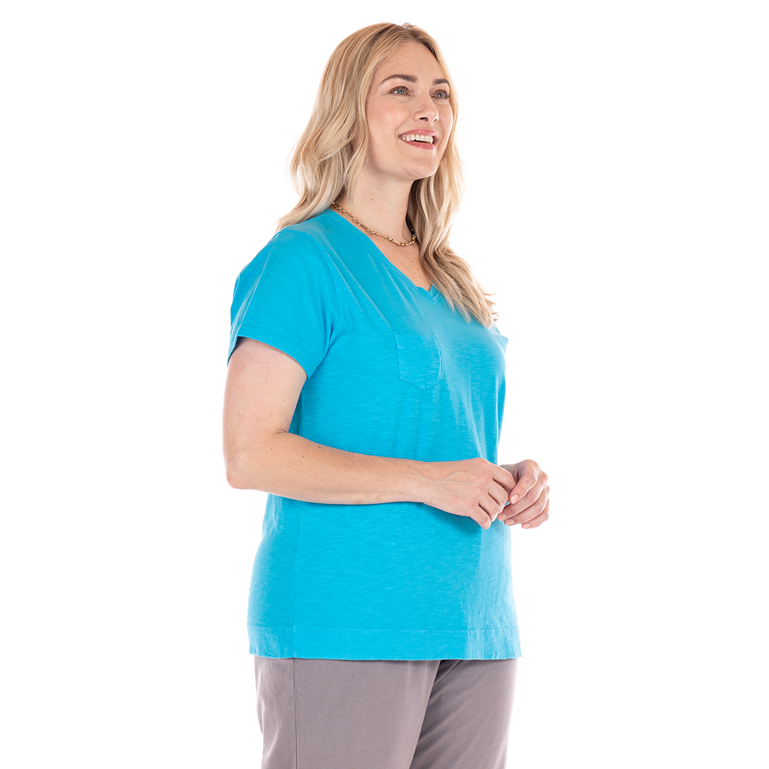 A woman with long blonde hair wears the Heritage Logo Fem T in bright blue from Fresh Produce Clothes and gray pants, standing and smiling against a white background.