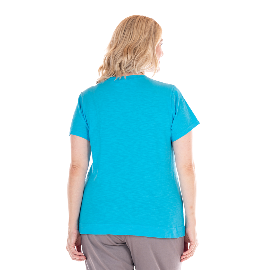 A person with shoulder-length blonde hair is standing with their back to the camera, wearing a bright blue Fresh Produce Clothes Heritage Logo Fem T | Plus Size and light gray pants against a white background.