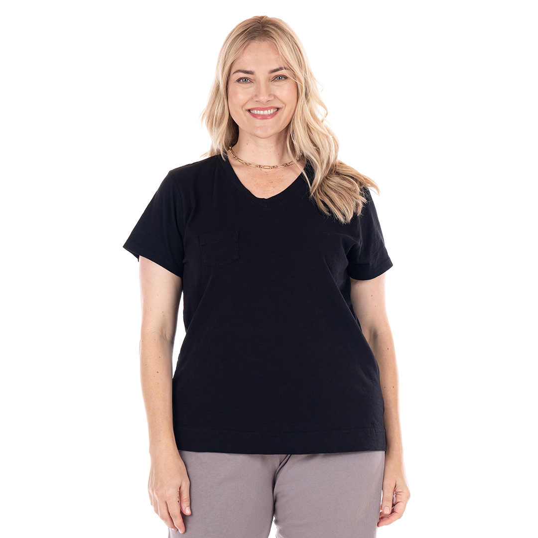 A woman with long blonde hair smiles while wearing a black Heritage Logo Fem T cotton slub tee and light gray pants, standing against a white background.