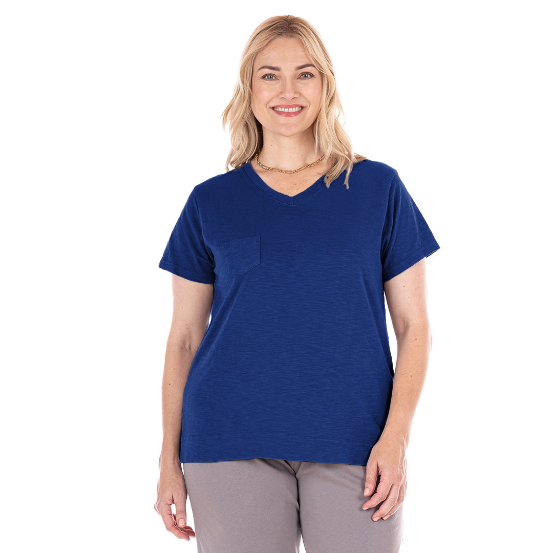 A smiling woman with shoulder-length blonde hair is wearing the Fresh Produce Clothes Heritage Logo Fem T | Plus Size, a blue V-neck T-shirt with a pocket, paired with light gray pants, standing against a white background.